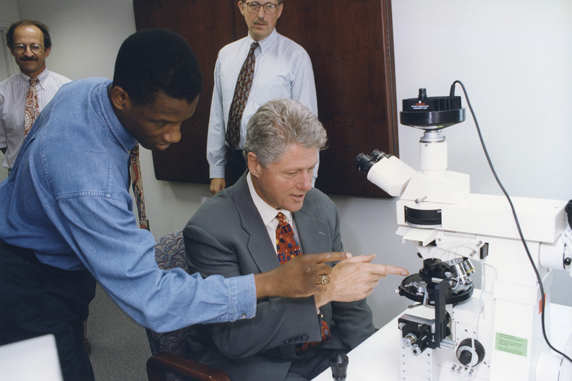 Rodney Wiltshire, former NHGRI fellow, shows President Bill Clinton how to do a chromosome micro-dissection. Former NIH Director Harold Varmus and current NIH Director Francis Collins are in the background.